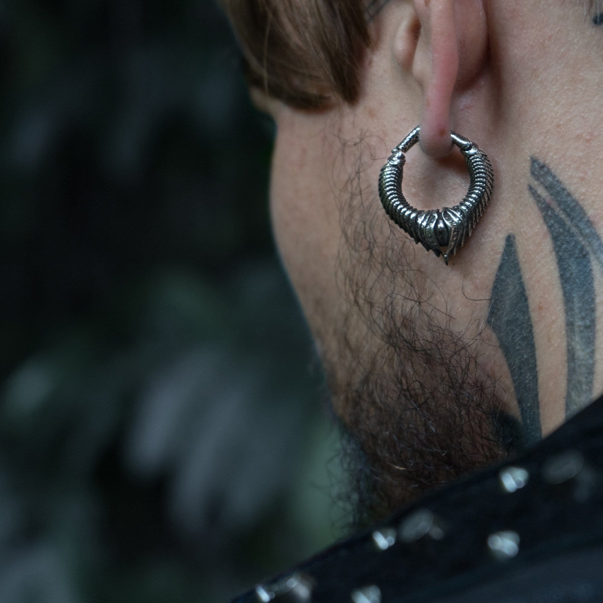 Close-up of the side profile of a person's head, revealing a silver MARVDASHT Small Biomechanical Clicker Ear Weight by Kaya Movement in their ear and a tattoo with pointed shapes on the neck. The background is blurred with shades of green and grey, suggesting an outdoor setting adorned with post-apocalyptic body jewelry.